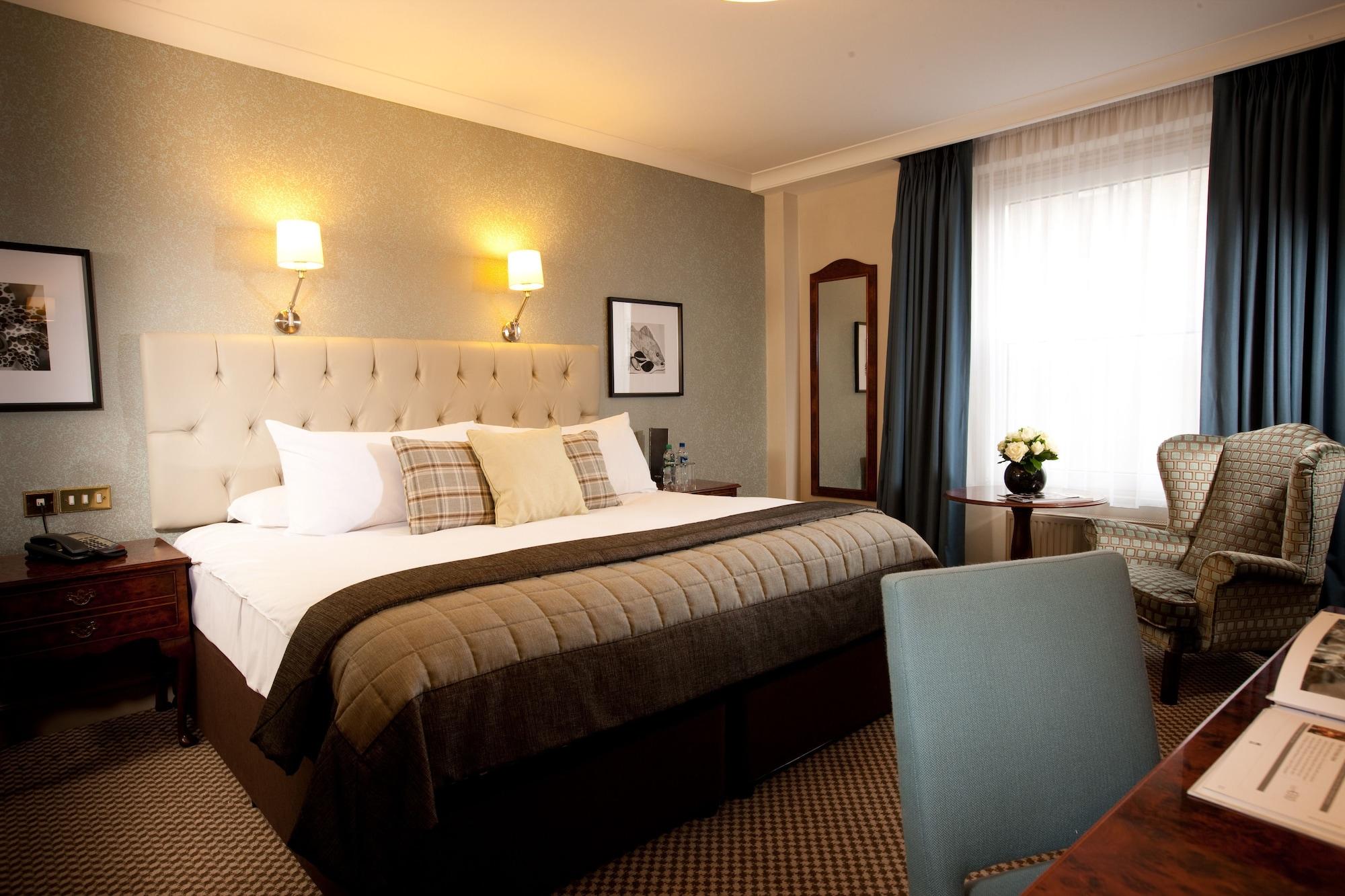Hotel The Grand Hove Esterno foto The photo shows a well-decorated hotel room. It features a large bed with a light-colored headboard and several decorative pillows. There are bedside lamps mounted on the walls next to the bed. The room has a window with light-colored drapes, allowin