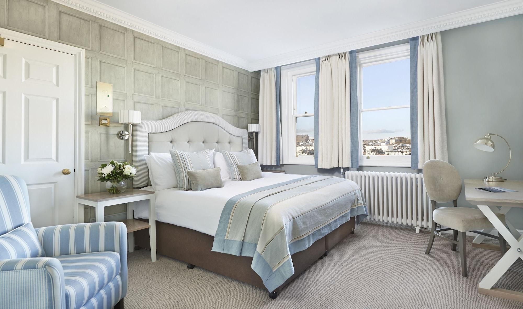Hotel The Grand Hove Esterno foto The image shows a well-decorated hotel room featuring a double bed with a light-colored headboard and soft bedding, complemented by decorative pillows. The walls have a subtle textured design, and there are large windows that allow natural light to f
