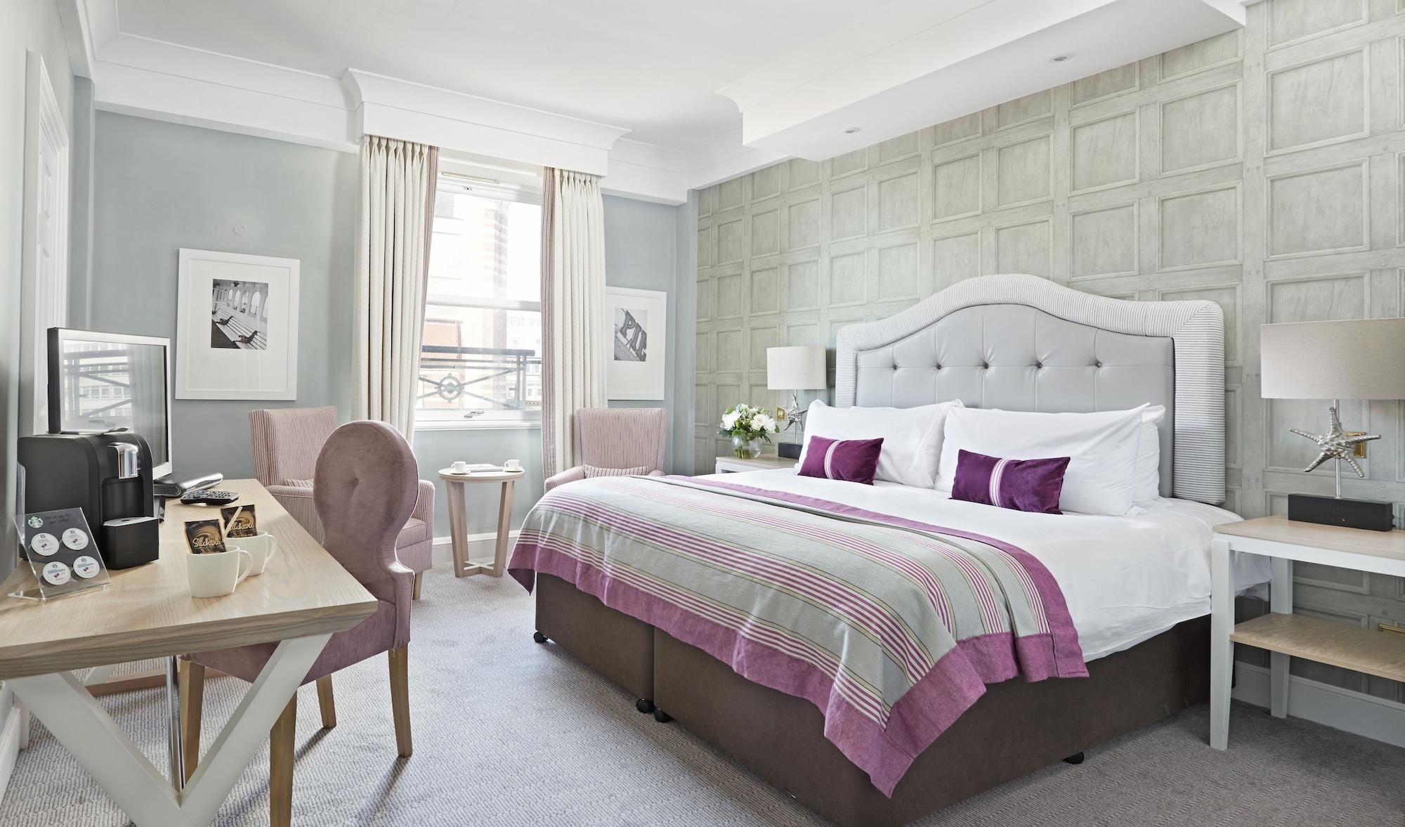 Hotel The Grand Hove Esterno foto The photo shows a stylish and modern hotel room. It features a large bed with a light gray upholstered headboard and white bedding accented with purple pillows. The wall behind the bed has a textured design in a light color. There are two framed pict