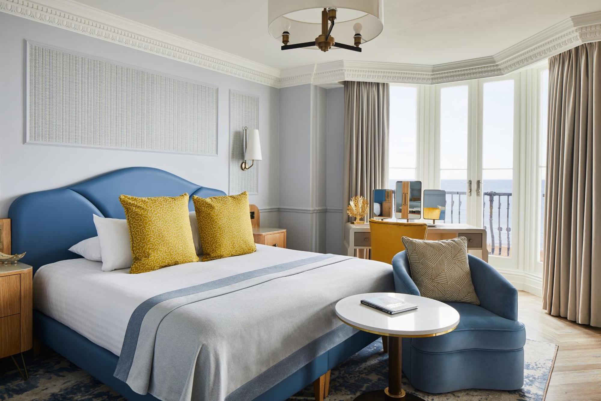 Hotel The Grand Hove Esterno foto The image shows a well-decorated hotel room featuring a large bed with a blue headboard and coordinating bedding. There are two yellow decorative pillows on the bed, adding a pop of color. A small circular table is placed next to a blue armchair, cre