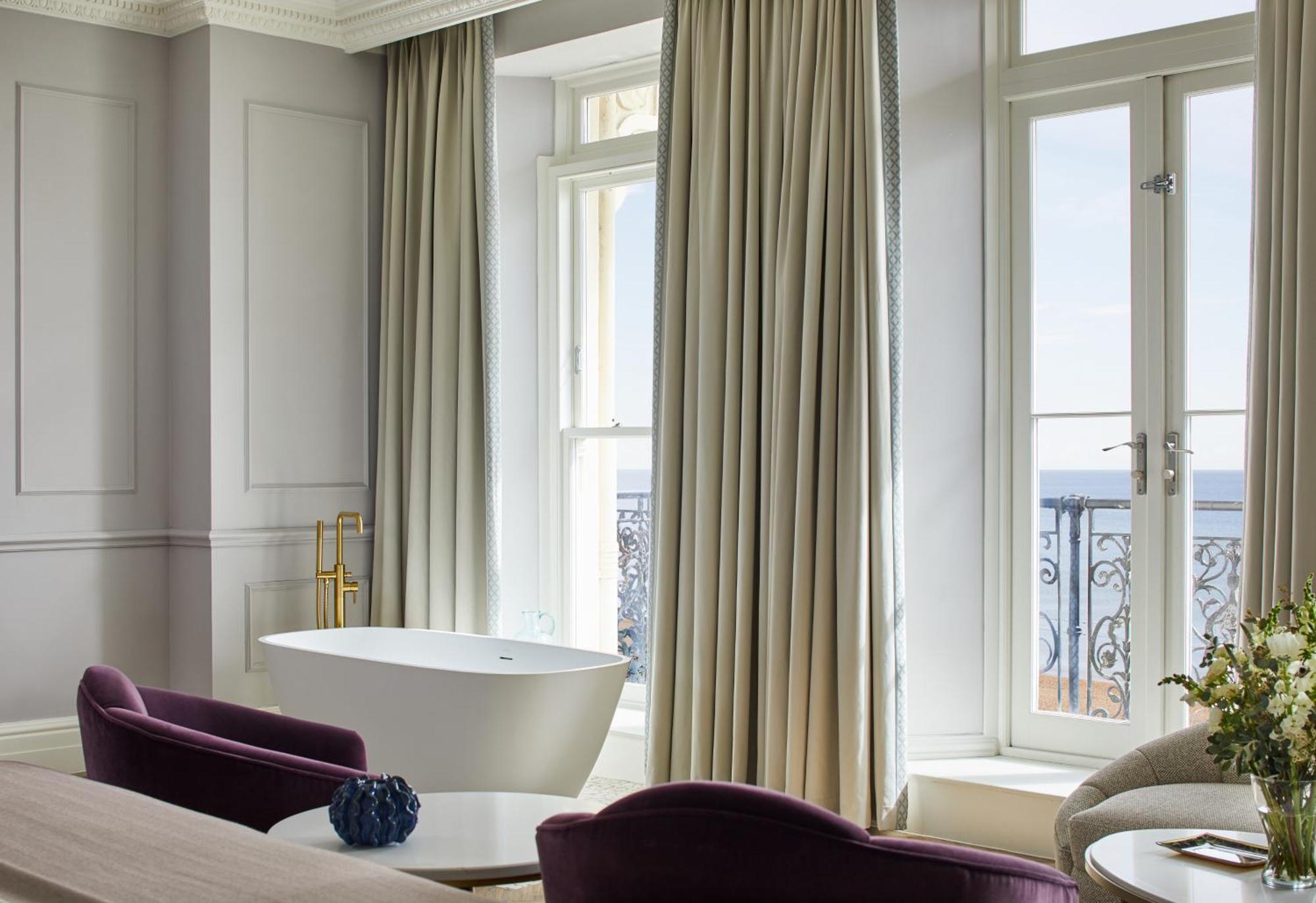 Hotel The Grand Hove Esterno foto The photo depicts a stylish room with large windows allowing natural light to enter. There is a freestanding bathtub near the window, and elegant, long curtains frame the windows. Two plush purple chairs are positioned near a wooden table, creating a