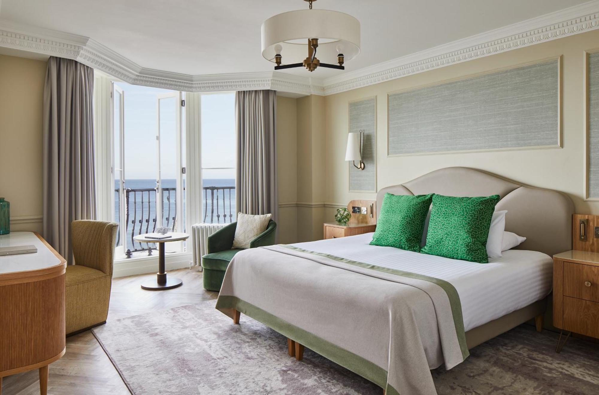 Hotel The Grand Hove Esterno foto The photo shows a well-appointed hotel room featuring a large bed with light-colored bedding and two green decorative pillows. There is a bedside table with a lamp on either side of the bed. The room has two windows that let in natural light, and the