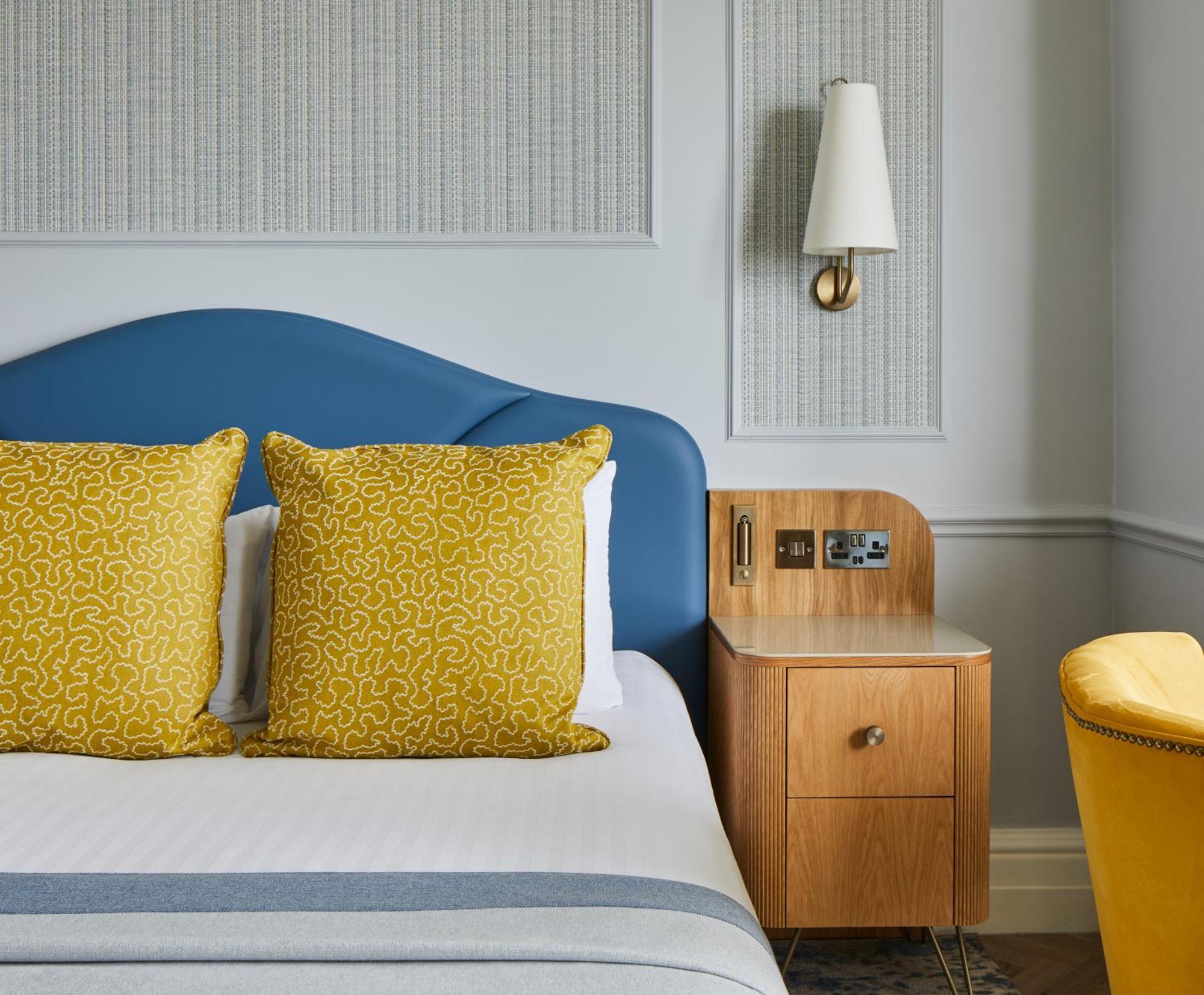 Hotel The Grand Hove Esterno foto The photo shows a bedroom with a modern and stylish design. There is a bed with a curved blue headboard and two yellow decorative pillows on it. Next to the bed, there is a wooden nightstand featuring an electrical outlet and a drawer. On the wall ab