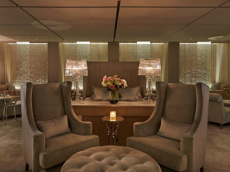 Hotel The Grand Hove Esterno foto The photo depicts a stylish and elegant interior of a lounge or seating area. In the foreground, there are two plush armchairs with tall backs, upholstered in a soft fabric, positioned on either side of a round, tufted coffee table. A small candle si